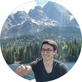 Person smiling while sitting by a lake with pine trees and mountains in the background on a sunny day.