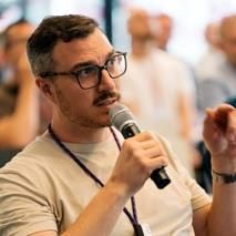 Man holding a microphone and speaking at an event, wearing glasses and a light-coloured t-shirt, with an attentive audience blurred in the background.
