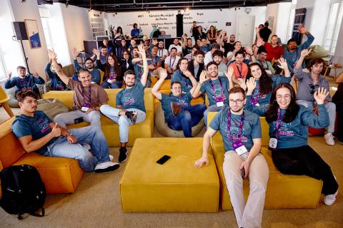 A large group of smiling participants sit on colorful couches and beanbags in a bright, modern room, many raising their hands toward the camera. Most are wearing matching teal hackathon T-shirts and conference badges, suggesting a friendly, collaborative event. The atmosphere is lively and energetic, with people chatting and engaging in the background.