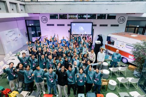 A large group of people wearing matching teal sweatshirts pose together in a bright, open indoor space, many smiling, raising their fists, or holding up number-one gestures in celebration. The group stands in front of a projection screen displaying a hackathon logo, with colorful chairs, tables, and a red-and-white camper van visible in the background. The setting has a lively and energetic atmosphere, suggesting the conclusion or celebration of a tech event or competition.