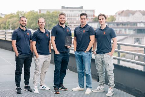 Five men stand outdoors on a modern walkway with buildings and trees in the background, smiling at the camera. They are all wearing matching navy-blue polo shirts with an orange logo, suggesting they are part of the same team or company. One person in the center holds a small chalkboard sign that reads “Team G6 #ByHackathon.” The group appears relaxed and professional, posing together in a confident, team-oriented manner.