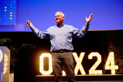 Man in blue shirt and brown pants presenting on a conference stage with raised hands