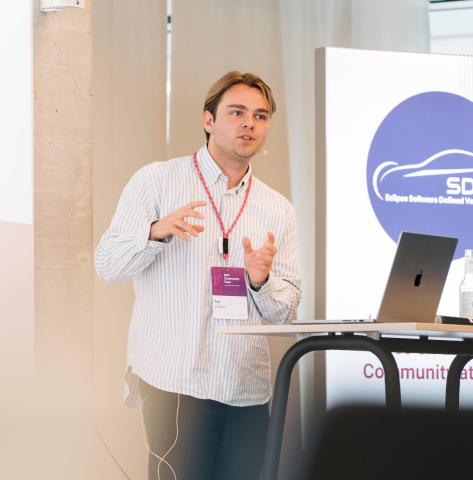 man with a conference badge standing in front of an open laptop, giving a talk