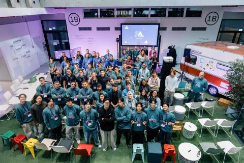 Large group photo of participants at an Eclipse SDV hackathon event, standing together in a bright indoor venue. Many attendees wear matching Eclipse SDV hackathon sweatshirts and badges. A presentation screen and event branding are visible in the background, along with seating areas and a retro-style camper van display.