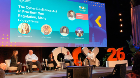Conference panel on stage discussing the Cyber Resilience Act, with five speakers seated and a large screen behind them displaying the session title, moderator Juan Rico, and speakers Maika Föhrenbach, Johan Klykens, Lola Fernandez, and Mike Milinkovich at Open Community Experience 2026.