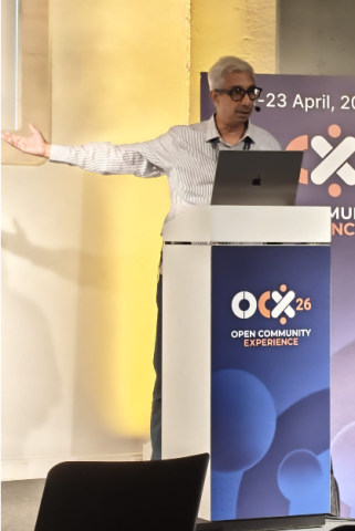 Manoj Pallat (IBM) Conference speaker presenting at Open Community Experience 2026, standing behind a podium with a laptop and gesturing towards a slide off screen.