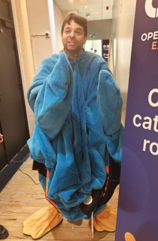 Matthews Khouzam wearing the OCX blue mascot costume with oversized sleeves and orange feet, standing in a hallway at a conference venue