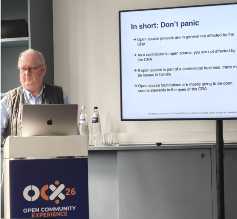 Speaker presenting at Open Community Experience 2026, standing behind a podium with a laptop, next to a slide titled ‘In short: Don’t panic’ explaining that open source projects and contributors are generally not affected by the Cyber Resilience Act.