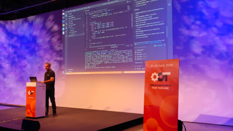 Jonas Helming giving a live coding demonstration on stage at Open Community for Tooling 2026, with a large screen showing a code editor interface and terminal output behind him.